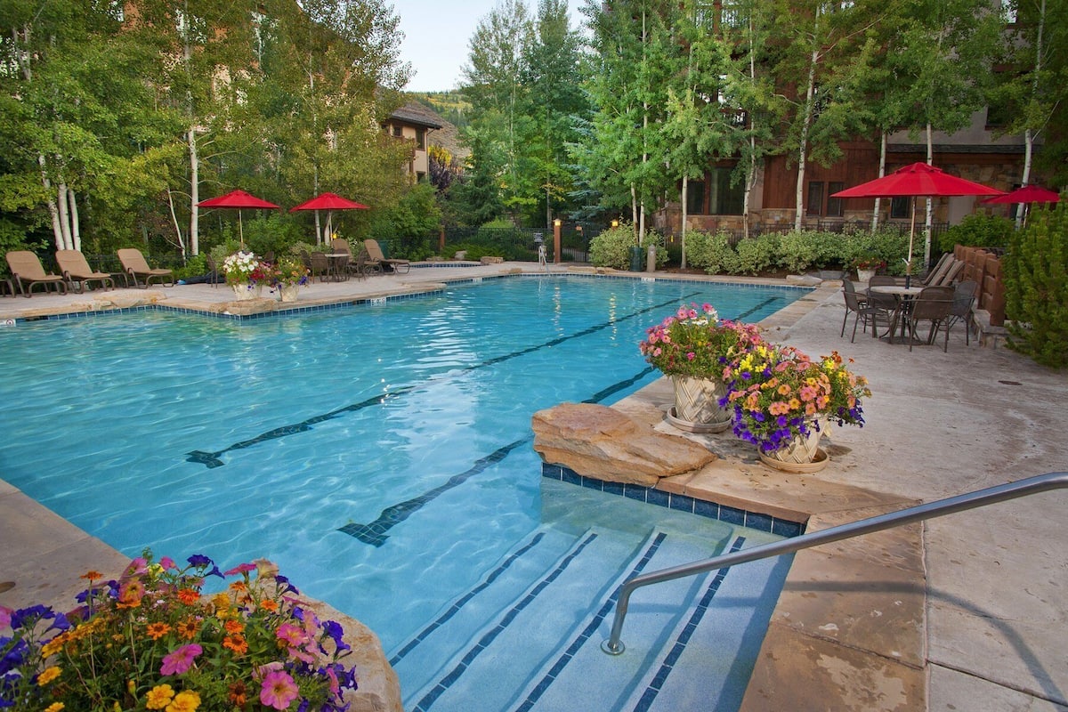 A swimming pool with a stone border, lounge chairs, and red umbrellas.