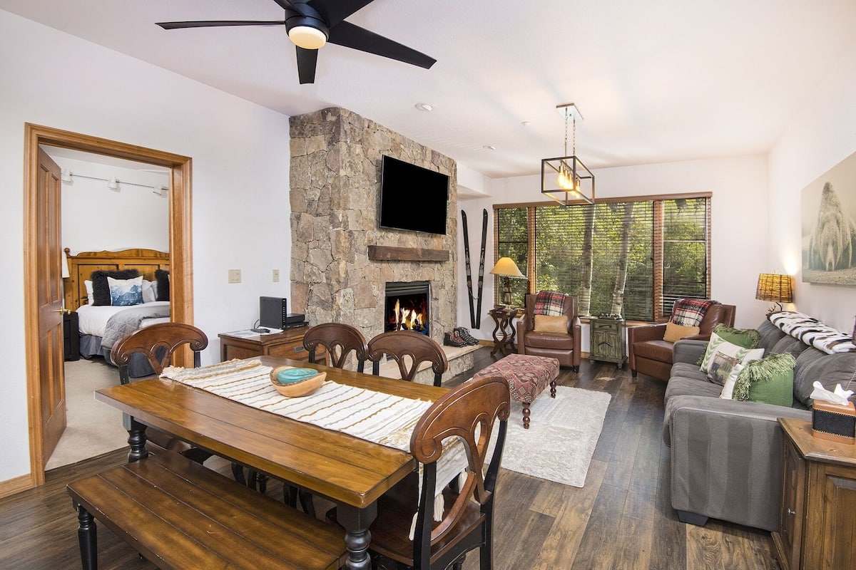 A spacious living room with a stone fireplace, a dining area with wooden furniture, and a large window providing a view of the outdoors.