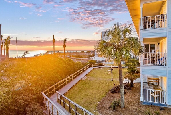 A beachfront property with a wooden deck, palm trees, and a view of the ocean at sunset.