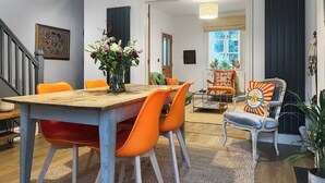 Uma sala de jantar com uma mesa de madeira e cadeiras laranja, um vaso com flores e vista para uma sala de estar com uma poltrona e uma planta.
