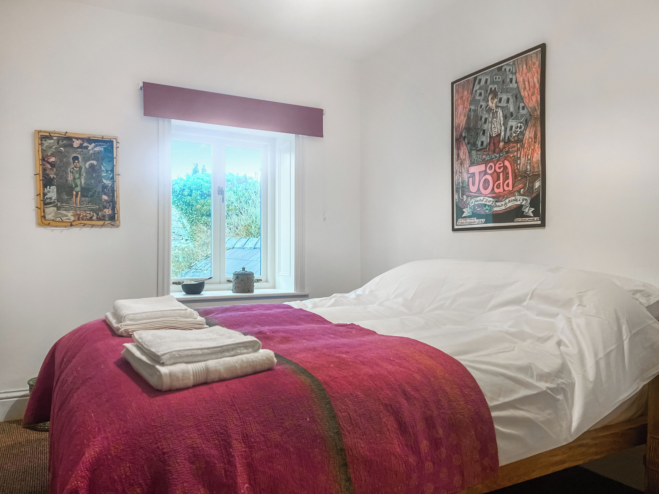 Een slaapkamer met een bed, een raam met uitzicht op groen en twee kunstwerken in lijsten aan de wanden.