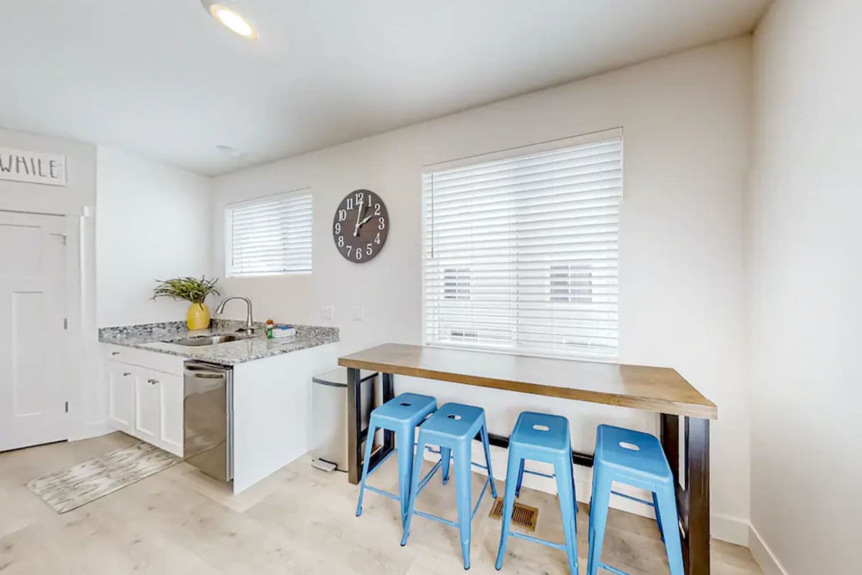 A modern kitchen with a central island, bar stools, a clock, and a window with blinds.