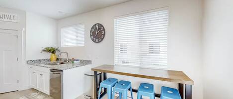 A modern kitchen with a central island, bar stools, a clock, and a window with blinds.
