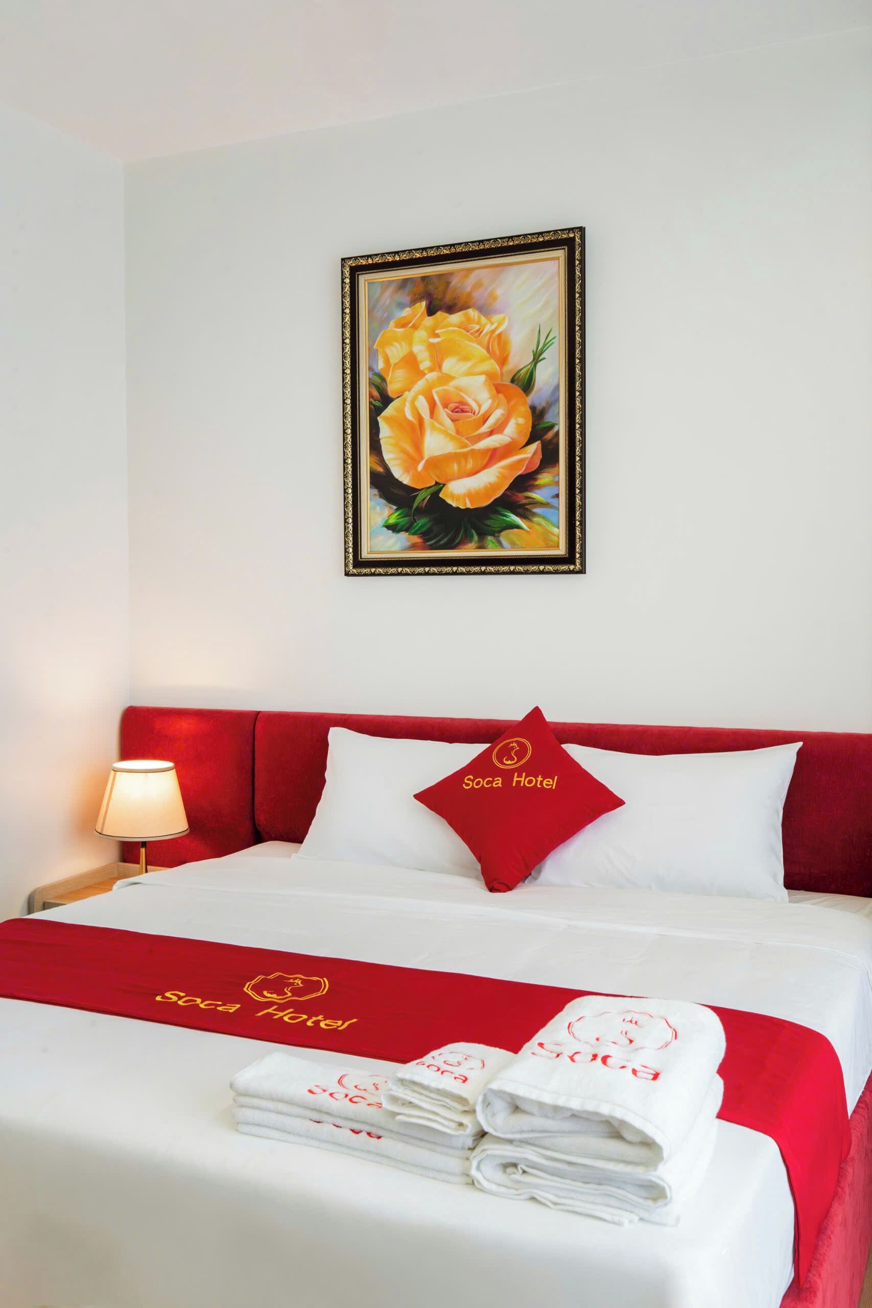 A hotel room with a red headboard, a painting of yellow roses, a pillow with the hotel's logo, and a bed with white linens and red towels.