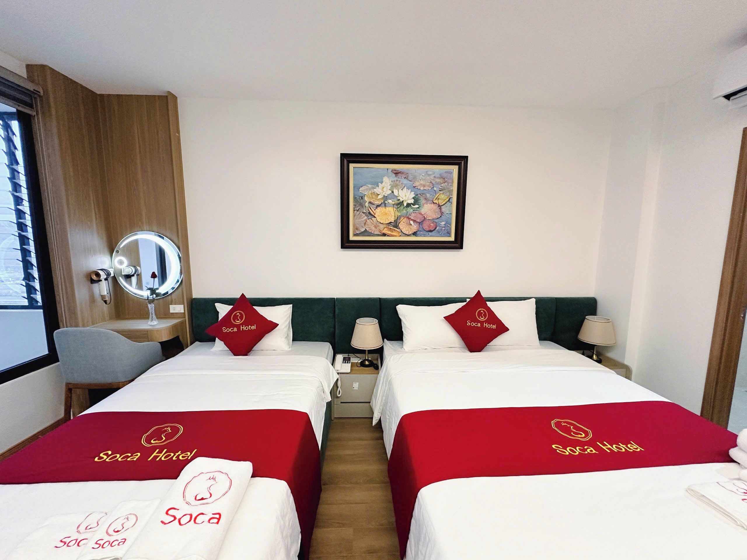 A hotel room with two beds, each with a red bedspread and pillows featuring the hotel's logo. There is a bedside table with a lamp, a small table with a phone, and a framed painting on the wall.