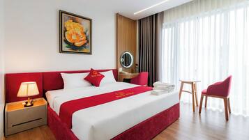A hotel room with a large bed, a bedside table with a lamp, a red chair, a small round table, and a painting of a rose on the wall.