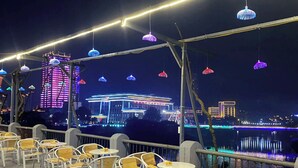 Een terras met stoelen en tafels, met uitzicht op een rivier met verlichte gebouwen op de achtergrond.