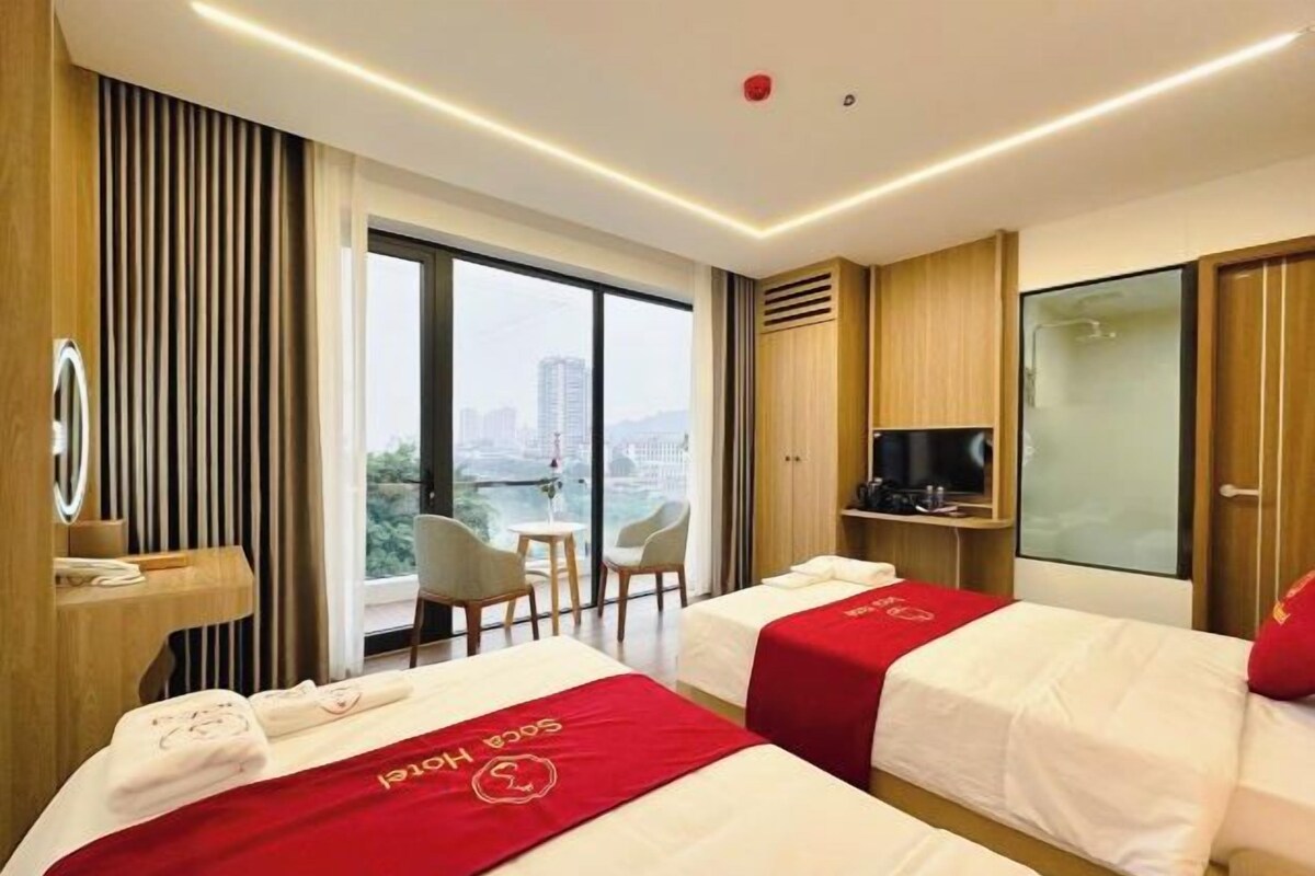 Ein Hotelzimmer mit zwei Betten, einem Fernseher, einem Schreibtisch und Blick auf die Stadt.