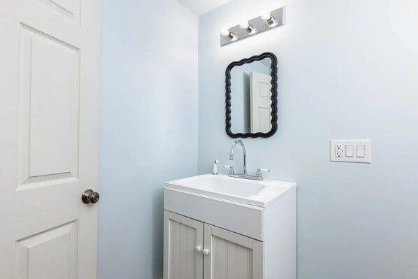 A bathroom with a white door, a white sink with a mirror above it, and a light switch on the wall. - Meredith Town & Trail Cottage - walkable to water/downtown, dogs ok! (Meredith)