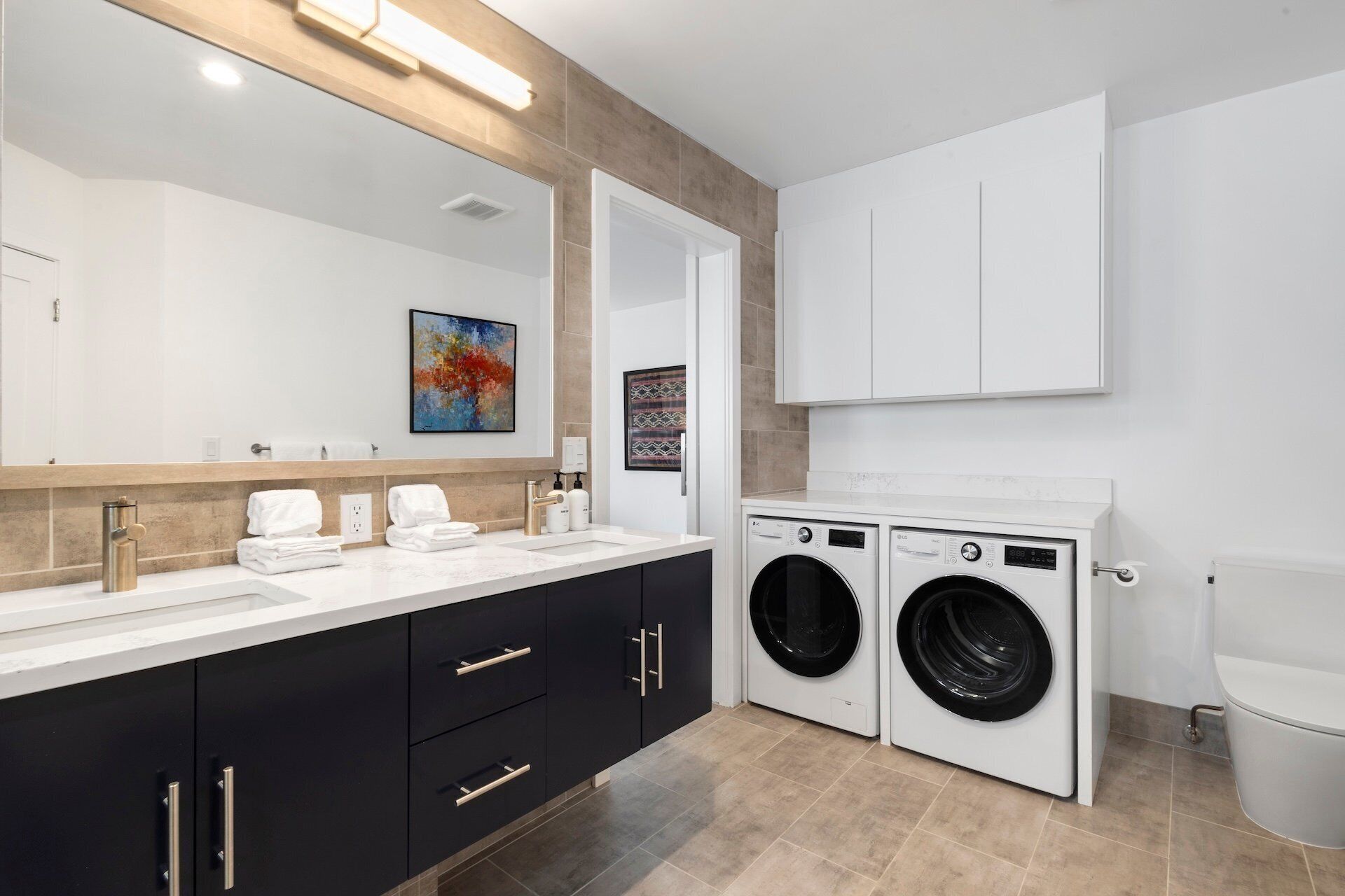 A modern bathroom with a double sink vanity, a large mirror, a washing machine, and a toilet.