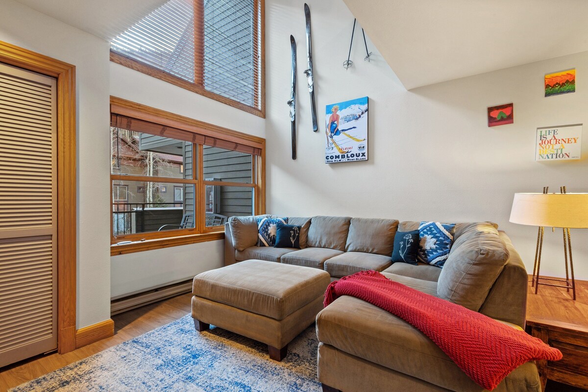 A living room with a sectional sofa, ottoman, and a window with blinds.