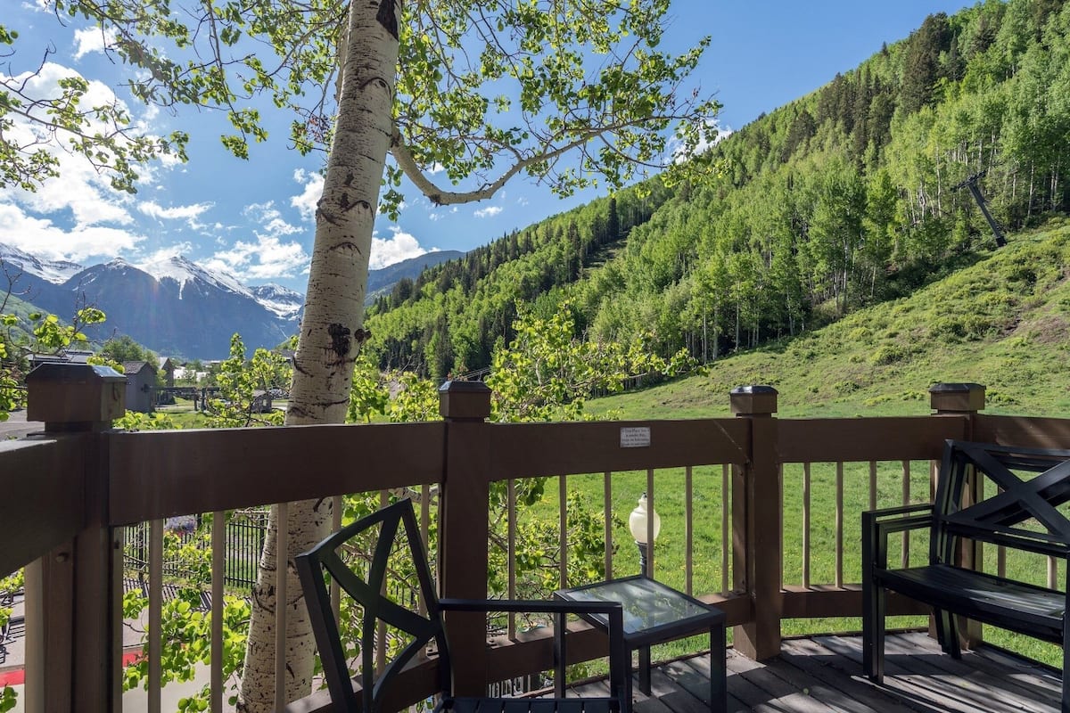 A wooden deck with a glass table and benches overlooking a lush green valley and mountains.
