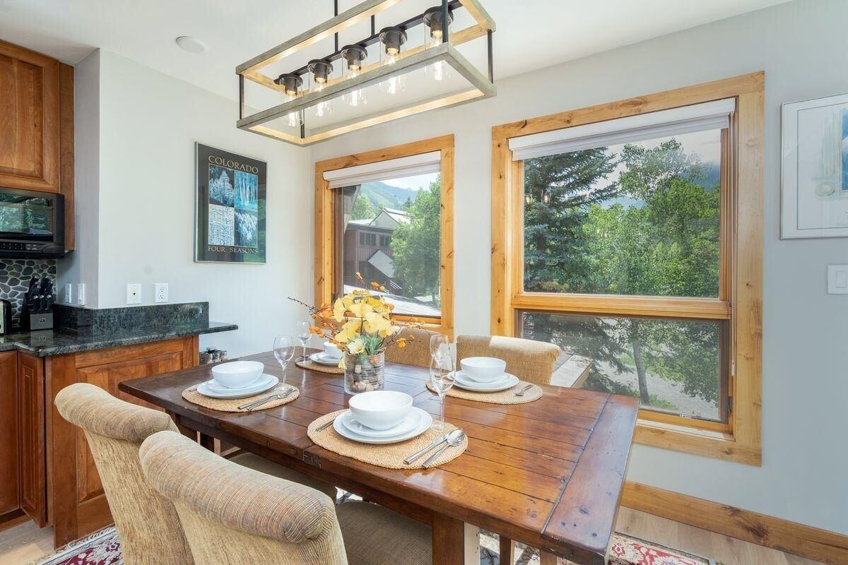 A dining area with a wooden table set for four, large windows, and a kitchen with a stone backsplash.