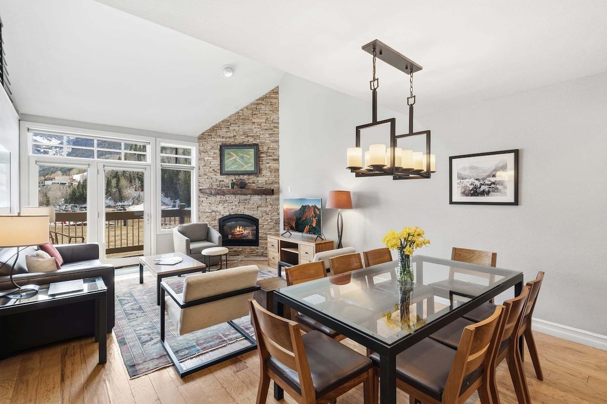 A modern living room with a fireplace, dining area, and large windows overlooking a snowy landscape.