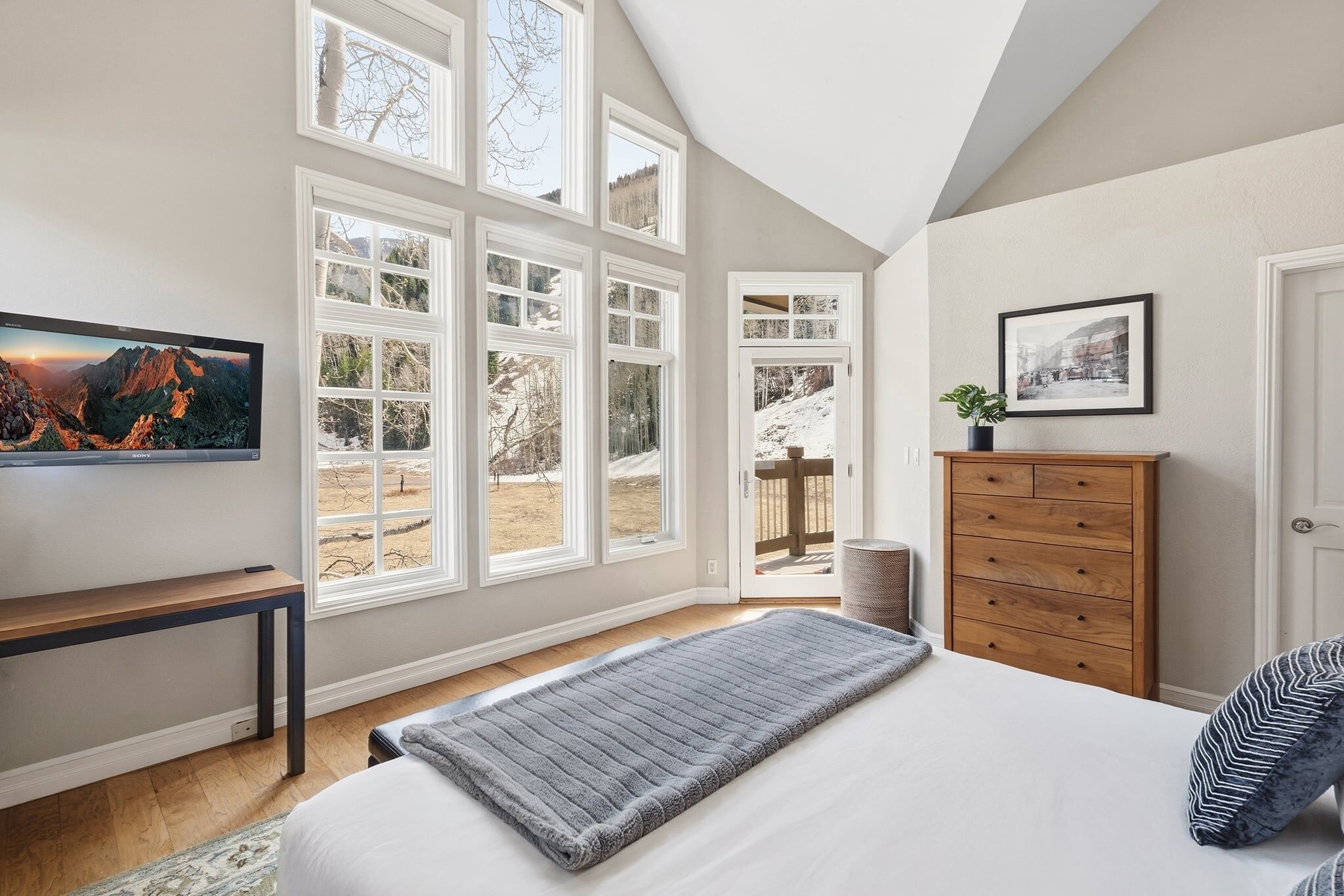 A bedroom with a bed, a TV, a wooden dresser, and large windows.
