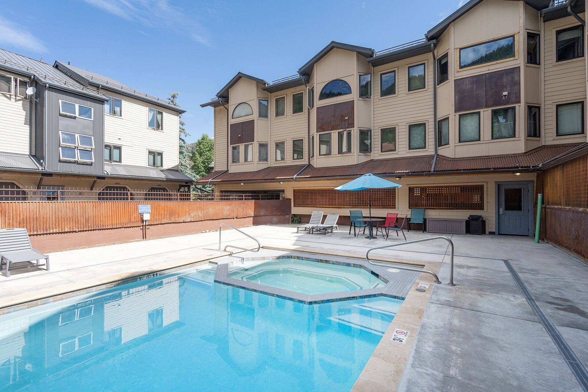A swimming pool with lounge chairs and umbrellas, surrounded by a fenced area with buildings in the background.
