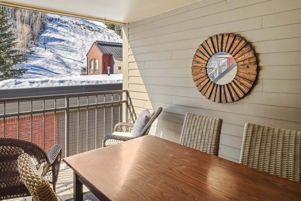 A dining area with a wooden table and wicker chairs, a round mirror on the wall, and a view of a snowy landscape through a window.