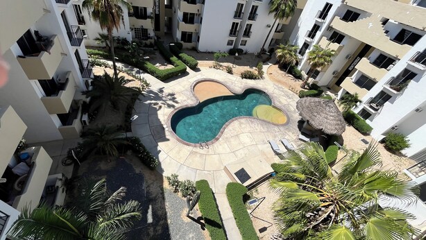 A multi-story residential building with a swimming pool, palm trees, and a thatched-roof structure.