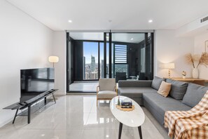 Ein modernes Wohnzimmer mit einem Flachbildfernseher, einer Couch, einem Couchtisch und Blick auf eine Stadtlandschaft durch große Fenster.