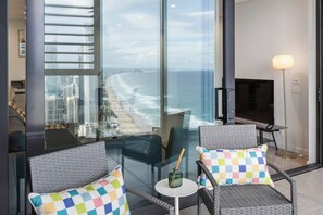 A modern living room with a large glass window overlooking a beach and cityscape. There are two wicker chairs with checkered cushions, a small round table with a vase, and a floor lamp. - Ocean Luxe Retreat 2  Kingbeds Free Parking (Surfers Paradise)
