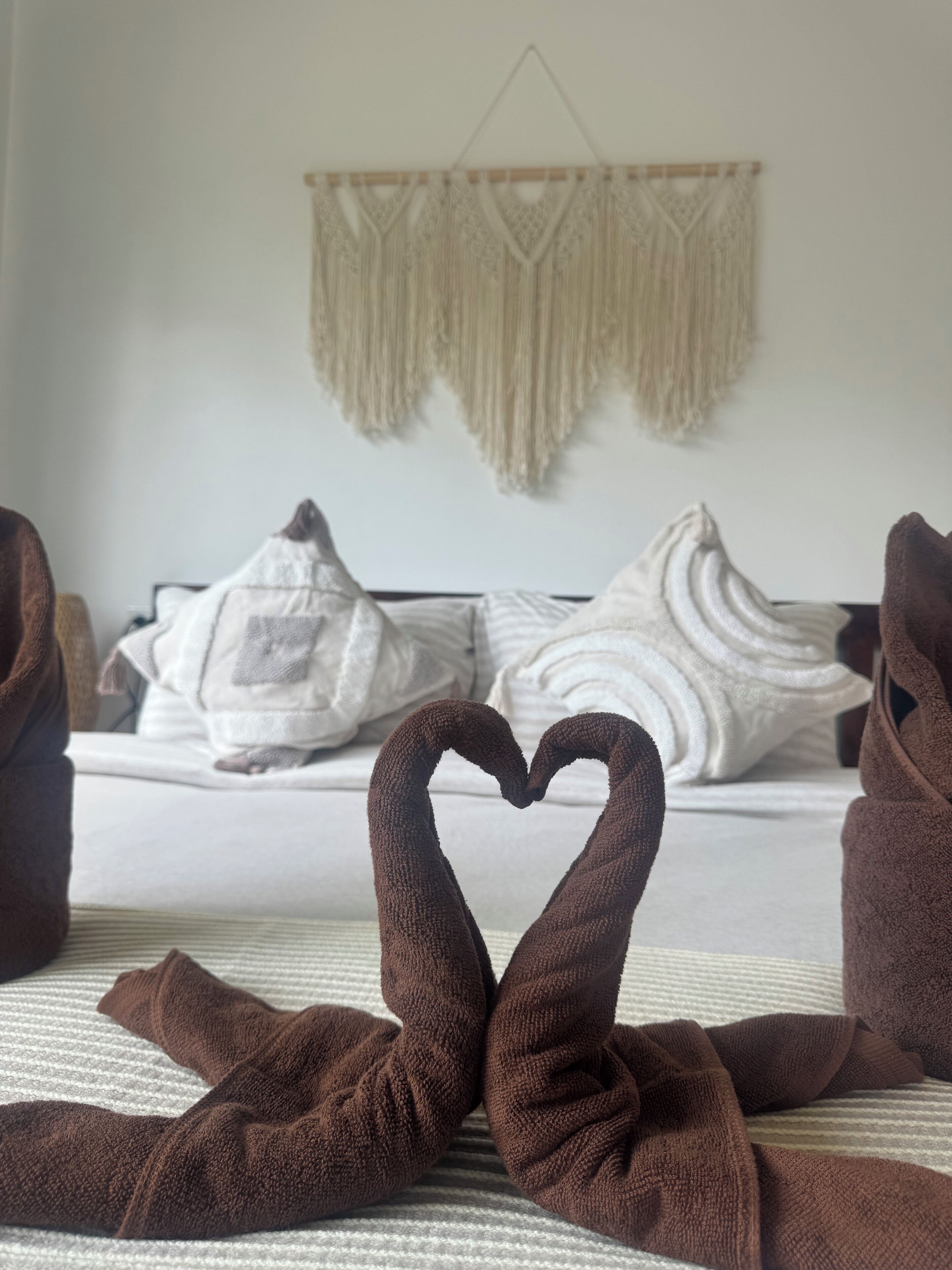 A bed with a heart-shaped towel arrangement, a macrame wall hanging, and a striped bedspread.