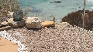 A coastal view with clear turquoise waters, a pebble beach, and a rocky hillside.