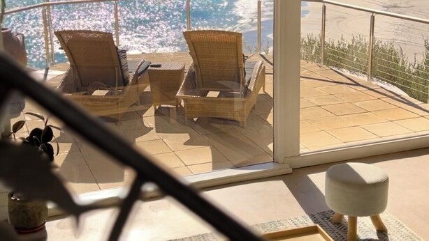 A balcony with wicker chairs and a table, overlooking a beach and ocean.