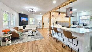A modern living room with a sofa, coffee table, and a TV. There is a kitchen area with a bar and stools. - Modern Rustic Retreat - Cozy 4-Bedroom ATL (Decatur)
