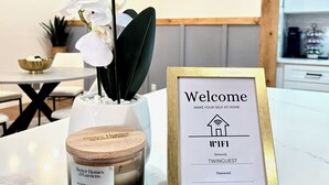 A kitchen area with a white orchid in a glass vase, a candle labeled 'Better Homes & Gardens', and a framed welcome sign with WiFi and guest details. - Modern Rustic Retreat - Cozy 4-Bedroom ATL (Decatur)