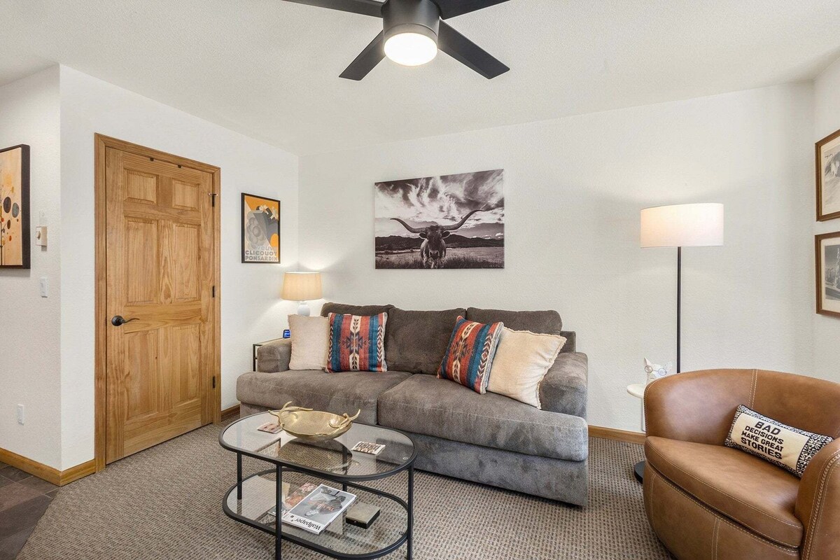 Un salon avec un canapé gris, un fauteuil en cuir marron, une table basse en verre, une porte en bois et un ventilateur de plafond.