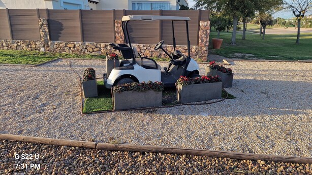 A golf cart parked in a gravel area with flower planters, a stone wall, and a building with a tiled roof in the background.
