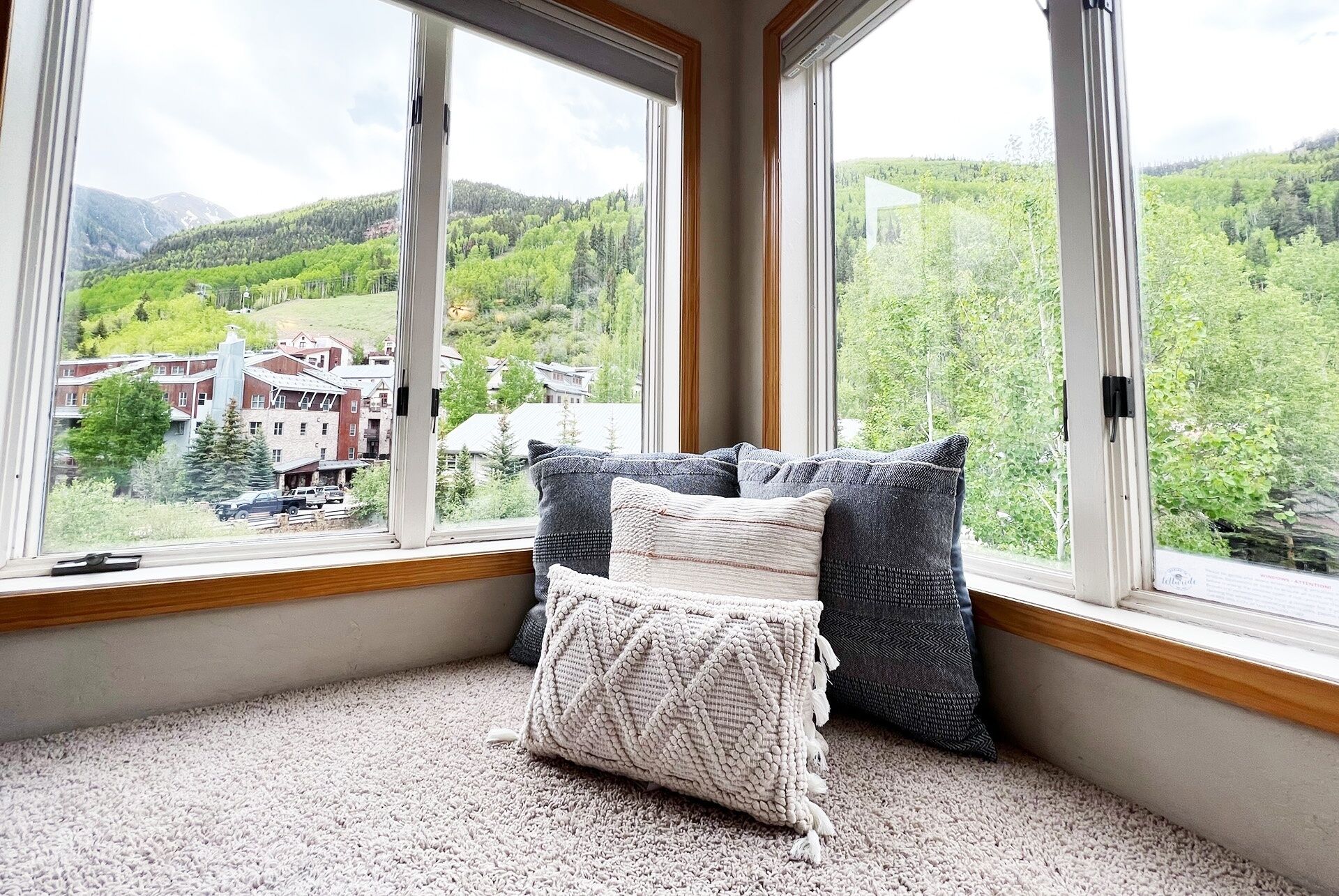 Une pièce dotée d’une grande fenêtre donnant sur un paysage verdoyant et des bâtiments. Le sol est recouvert d’un tapis et un coussin est posé sur le rebord de la fenêtre.