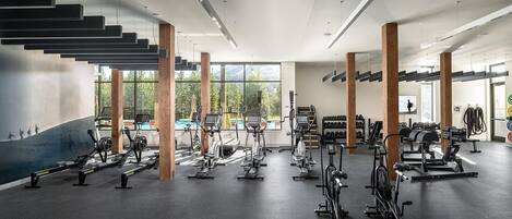 A modern gym with various cardio and strength training equipment, large windows, and a staircase.