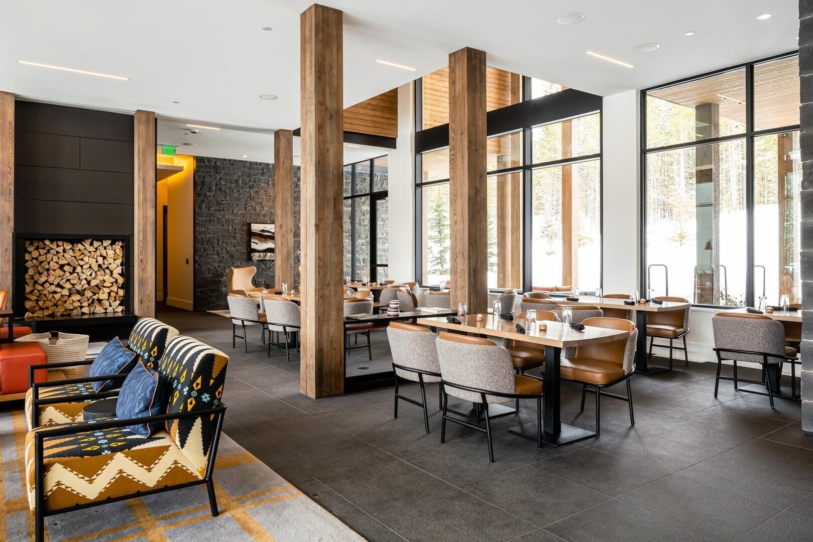 A modern dining area with high wooden pillars, large windows, and a stone wall with a fireplace.