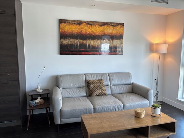 Ein modernes Wohnzimmer mit grauem Sofa, einem Holztisch, einer Stehlampe und einem Gemälde einer Waldszene.