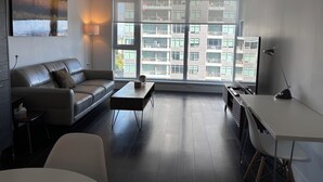 A modern living room with a sofa, coffee table, and desk. Large windows offer a view of a building with multiple floors. - Burnaby Metropolis Metrotown/1bedroom Condo/AC/Skytrain/Pool/Gym (Burnaby)