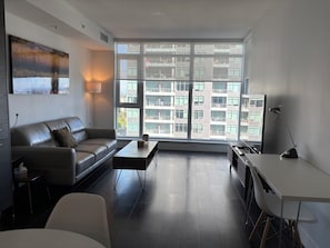Un salon moderne avec un canapé, une table basse et un bureau. De grandes fenêtres offrent une vue sur un immeuble à plusieurs étages.