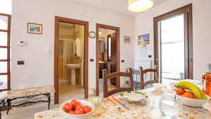 Un comedor con una mesa dispuesta para dos personas, un tazón de tomates, una jarra de agua y un cuenco con naranjas y plátanos.