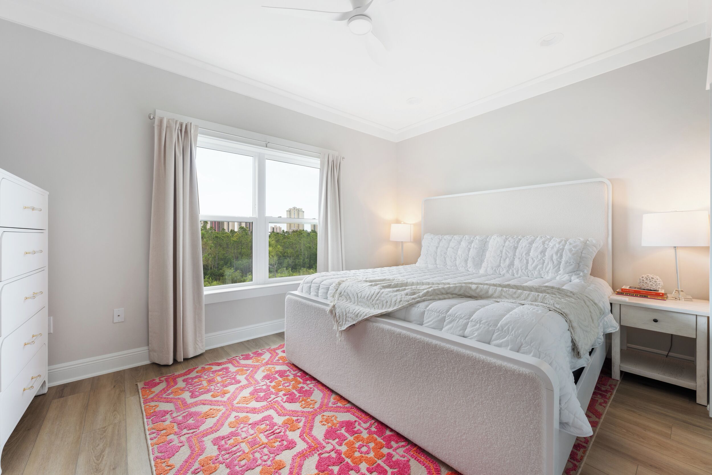 A bedroom with a bed, a nightstand, a dresser, a window with curtains, and a view of greenery outside.
