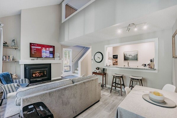 A modern living room with a fireplace, a TV mounted on the wall, a sofa, a dining area with a table and chairs, and a kitchen counter with a clock and a painting. - Glacier WA Mt Baker Vacation Rentals (Deming)