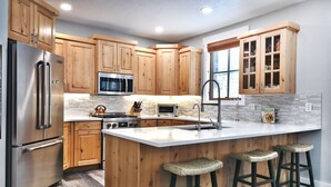 A modern kitchen with wooden cabinets, stainless steel appliances, a large island with bar stools, and a window with blinds. - Park City Ski Resort - Town Pointe Condos (Park City)