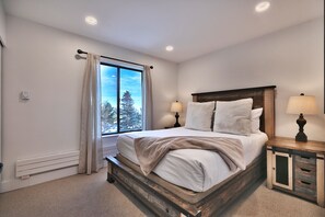 Ein Schlafzimmer mit großem Fenster, einem Bett mit weißer Bettwäsche, zwei Nachttischlampen und einem Nachttisch aus Holz.