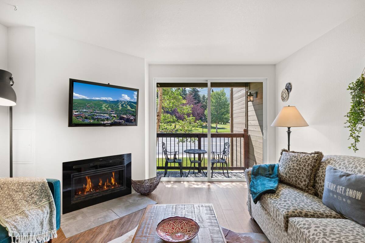 A living room with a fireplace, a television mounted on the wall, a sofa, a coffee table, and a balcony with outdoor seating.
