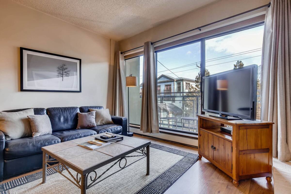 A living room with a black leather sofa, a coffee table, a wooden TV stand, and a large window with curtains.