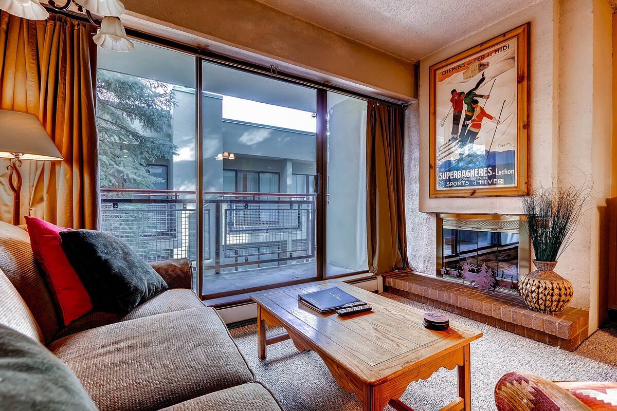 A living room with a sofa, a coffee table, a fireplace, and a large window with curtains.