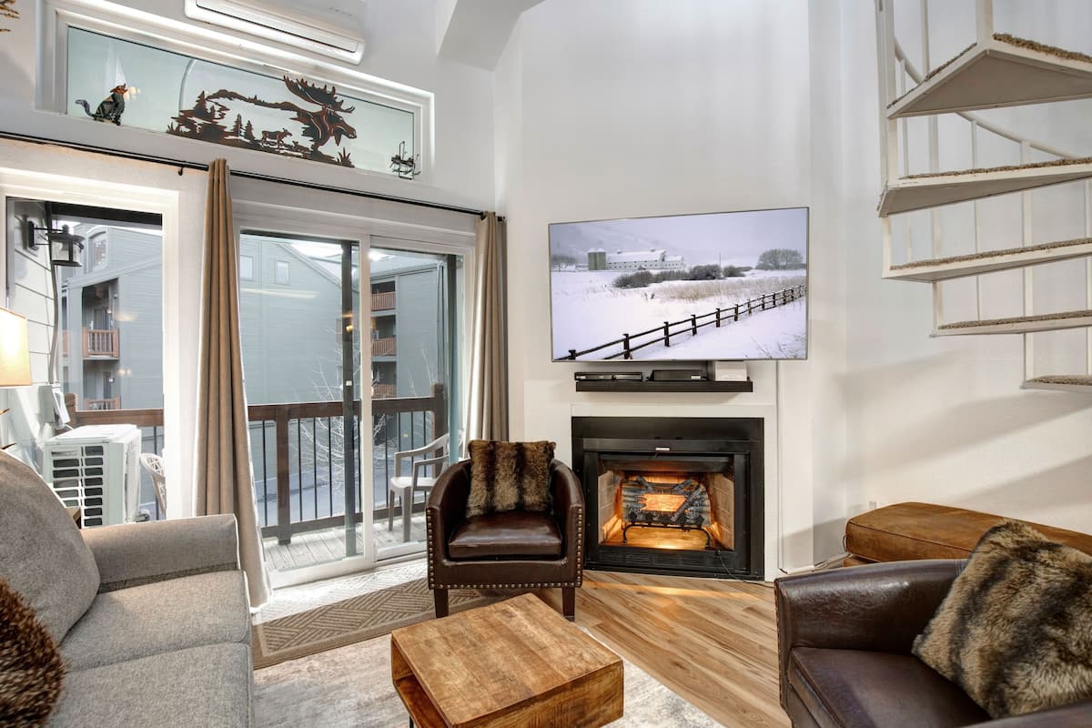 A cozy living room with a fireplace, a television mounted on the wall, and a large window overlooking a deck.