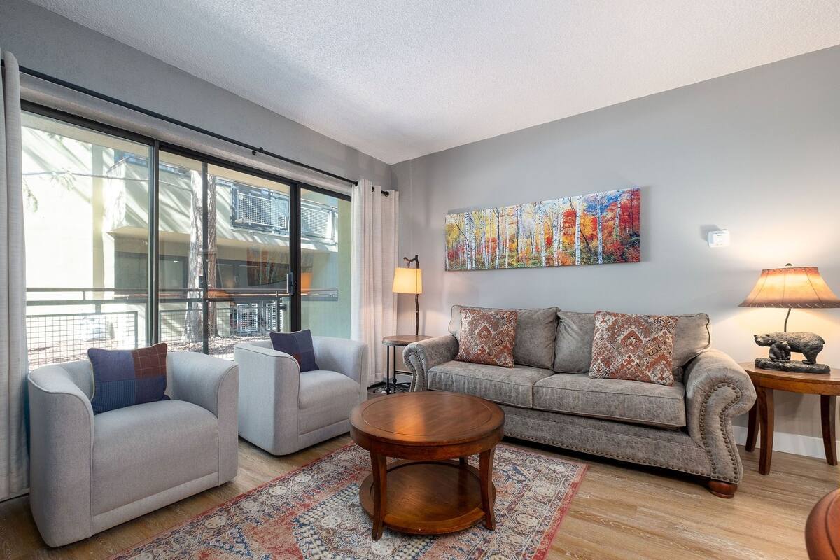 A modern living room with a large window, a sofa, two armchairs, a round wooden coffee table, and a painting on the wall.