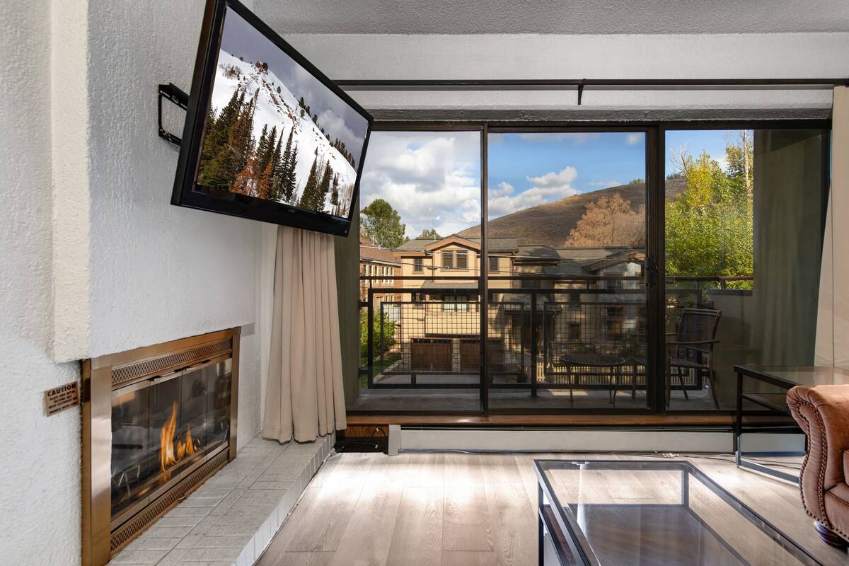 A modern living room with a fireplace, large sliding glass doors, a flat-screen TV, and a view of a residential area.