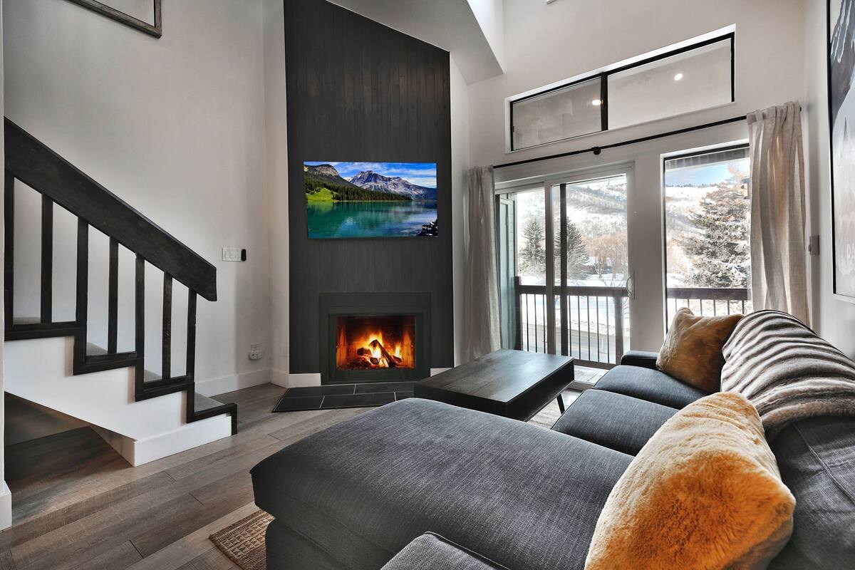 A modern living room with a fireplace, a large flat-screen TV, a sofa, and a window with a view of trees.