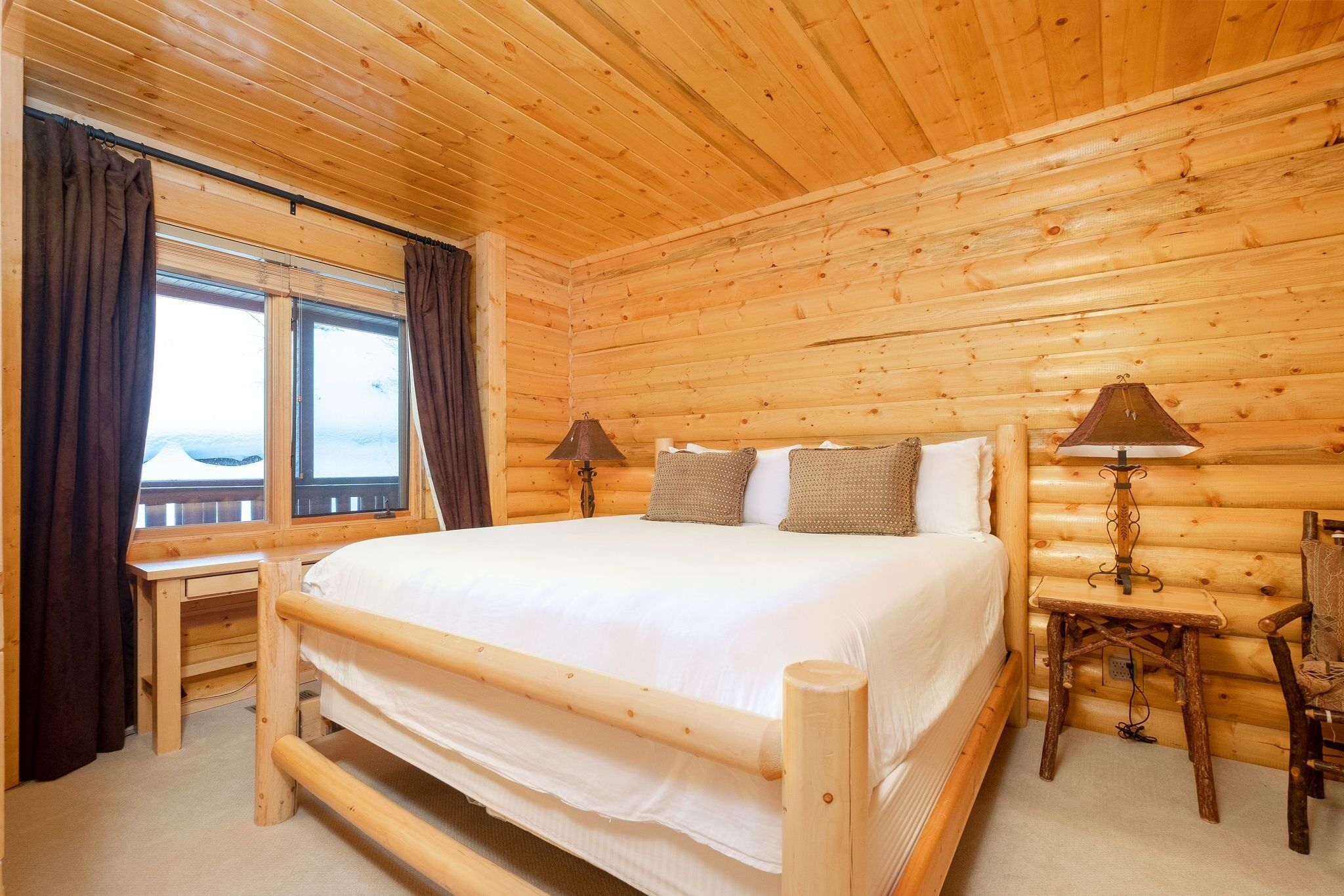 Une chambre dans une cabane en bois, avec un lit, deux lampes de chevet, une petite table et une fenêtre avec des rideaux.